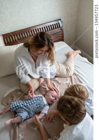 Mother and two children play with a newborn baby on a bed near a window, soft daylight, neutral colors, warm and caring family atmosphere 134957723