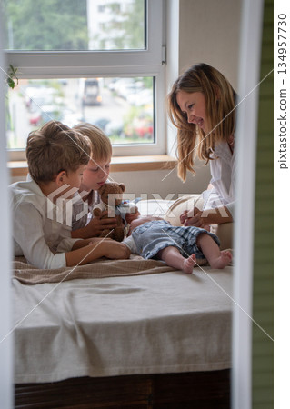 Mother and two children play with a newborn baby on a bed near a window, soft daylight, neutral colors, warm and caring family atmosphere 134957730