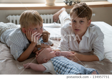 Three brothers lying together on a bed at home, older siblings gently interacting with a newborn baby in soft natural daylight Three brothers lying together on a bed at home, older siblings gently interacting with a newborn baby in soft natural daylight 134957731