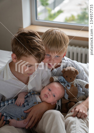 Three brothers sitting together on a bed at home, older siblings gently holding a newborn baby, soft natural light and calm interior 134957735