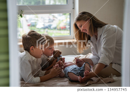 Mother and two children play with a newborn baby on a bed near a window, soft daylight, neutral colors, warm and caring family atmosphere 134957745