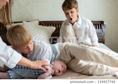 Mother and two young boys sit on bed with newborn baby while a mothers hand gently supports the infant, candid family moment Mother and two young boys sit on bed with newborn baby while a mothers hand gently supports the infant, candid family moment 134957748