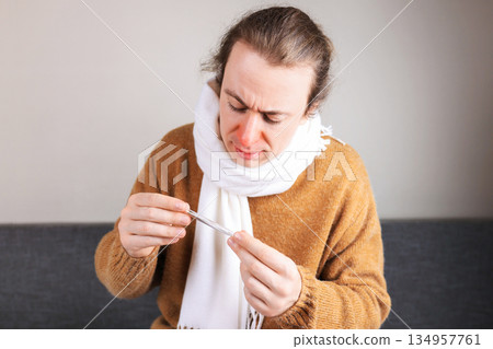 Sick man with red nose and scarf checking body temperature with thermometer. Flu and healthcare concept. High quality photo 134957761