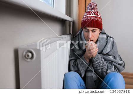 Frozen man in winter hat and blanket sitting near radiator and warming hands with breath at home. High quality photo 134957818