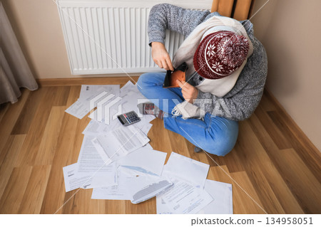 A man in winter clothes sits on the floor with utility bills and a calculator, checking his empty wallet near a radiator. High quality photo A man in winter clothes sits on the floor with utility bills and a calculator, checking his empty wallet near a radiator. High quality photo 134958051