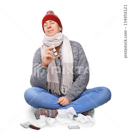 Sick man in winter hat holding nasal spray and feeling relief, sitting with tissues and pills isolated on white background. High quality photo 134958121