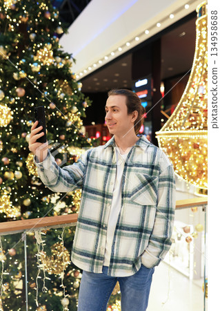 Young man taking selfie with smartphone in front of Christmas tree in decorated shopping mall. High quality photo 134958688