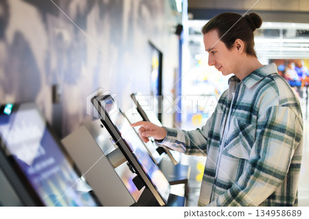 man using self-service kiosk with touchscreen. Guy selecting options on interactive terminal in cinema or mall. High quality photo 134958689