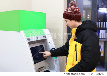 man in winter clothes using ATM with green mockup screen. Person withdrawing money at bank terminal. High quality photo man in winter clothes using ATM with green mockup screen. Person withdrawing money at bank terminal. High quality photo 134958692