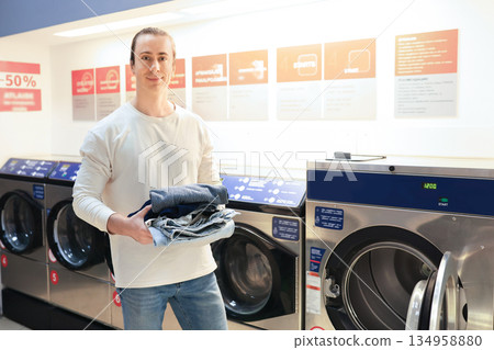 man holding stack of clean clothes in self-service laundromat with washing machines in background. High quality photo man holding stack of clean clothes in self-service laundromat with washing machines in background. High quality photo 134958880