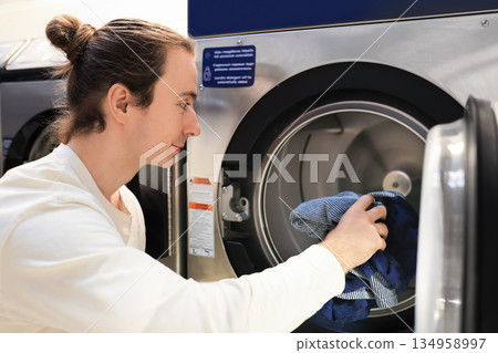 man with bun loading clothes into washing machine at public laundromat. High quality photo 134958997