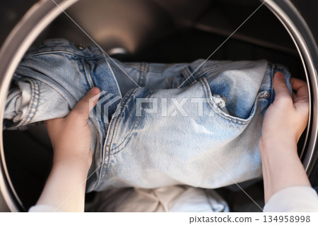 Hands loading blue jeans into washing machine drum close up view. High quality photo 134958998