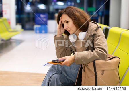 Thoughtful woman sitting at airport gate holding smartphone and passport, touching head Thoughtful woman sitting at airport gate holding smartphone and passport, touching head 134959566