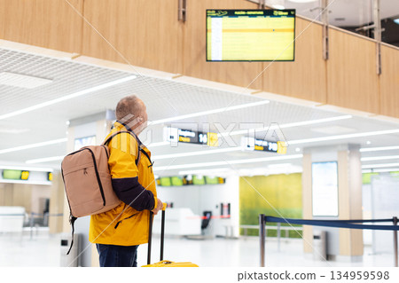 Middle aged man waiting at airport with suitcase, looking at departure board 134959598