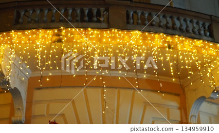 Hanging yellow and golden garlands of light bulbs swaying in wind against wall of an old building. New Year and Christmas. Festive street lighting. Decorating the facades of buildings. Winter spirit. Hanging yellow and golden garlands of light bulbs swaying in wind against wall of an old building. New Year and Christmas. Festive street lighting. Decorating the facades of buildings. Winter spirit. 134959910