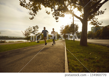 Two runners training on tree lined park path at sunrise, urban cardio workout 134960770