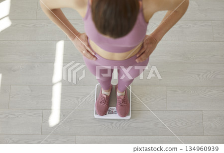 Portrait of a young sporty slim woman measuring her weight standing in scales at home. 134960939