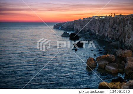 A breathtaking view of Malta's rugged stone coastline bathed in warm sunset hues, with golden light reflecting off the rocks and the tranquil Mediterranean Sea. 134961980