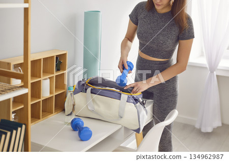 Sporty woman puts blue dumbbells in her spacious gym bag, preparing for her upcoming workout. 134962987