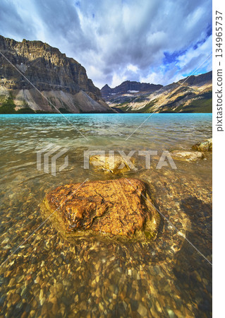 Bow Lake Crystal Water and Rocky Shore with Dramatic Mountain Range Under Cloudy Sky Bow Lake Crystal Water and Rocky Shore with Dramatic Mountain Range Under Cloudy Sky 134965737