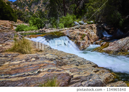 Waterfall Over Rocky Riverbank With Lush Greenery And Flowing Water In Sunlight Waterfall Over Rocky Riverbank With Lush Greenery And Flowing Water In Sunlight 134965848