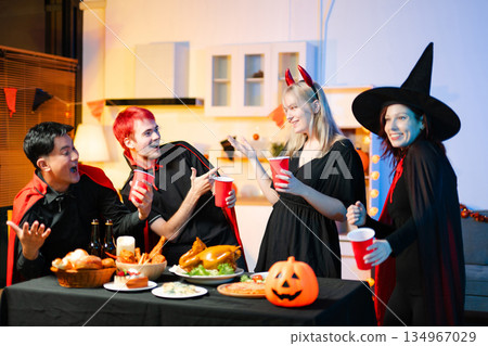 Friends in spooky Halloween costumes enjoy drinks and food at a festive indoor party with pumpkins, red cups, and laughter. Friends in spooky Halloween costumes enjoy drinks and food at a festive indoor party with pumpkins, red cups, and laughter. 134967029