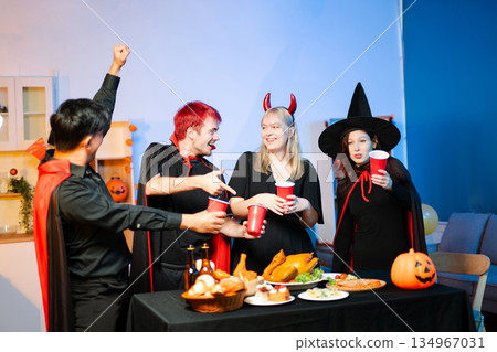 Friends in spooky Halloween costumes enjoy drinks and food at a festive indoor party with pumpkins, red cups, and laughter. Friends in spooky Halloween costumes enjoy drinks and food at a festive indoor party with pumpkins, red cups, and laughter. 134967031