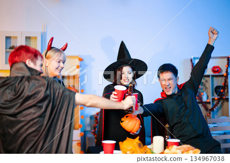 Group of young friends in Halloween costumes enjoy food and drinks at a cozy indoor party. with fun, spooky vibes, red cups, and pumpkin decor. Group of young friends in Halloween costumes enjoy food and drinks at a cozy indoor party. with fun, spooky vibes, red cups, and pumpkin decor. 134967038