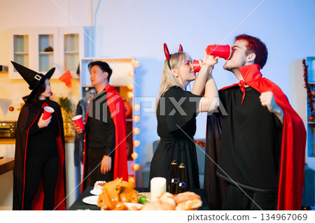 Group of young friends in Halloween costumes enjoy food and drinks at a cozy indoor party. with fun, spooky vibes, red cups, and pumpkin decor. Group of young friends in Halloween costumes enjoy food and drinks at a cozy indoor party. with fun, spooky vibes, red cups, and pumpkin decor. 134967069
