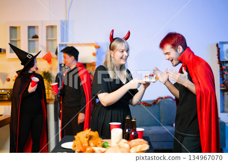 Group of young friends in Halloween costumes enjoy food and drinks at a cozy indoor party. with fun, spooky vibes, red cups, and pumpkin decor. Group of young friends in Halloween costumes enjoy food and drinks at a cozy indoor party. with fun, spooky vibes, red cups, and pumpkin decor. 134967070