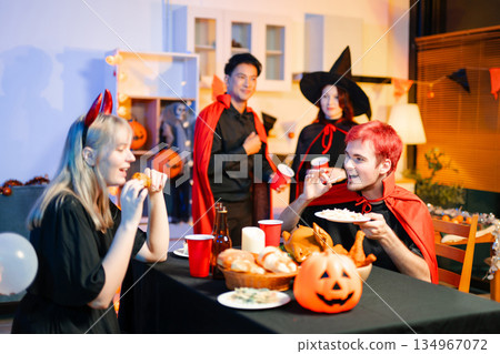 Group of young friends in Halloween costumes enjoy food and drinks at a cozy indoor party. with fun, spooky vibes, red cups, and pumpkin decor. 134967072