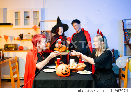 Young friends in Halloween costumes decorating pumpkin lanterns at a festive indoor party. Real people enjoying spooky vibes, red cups, snacks, and Halloween props. Young friends in Halloween costumes decorating pumpkin lanterns at a festive indoor party. Real people enjoying spooky vibes, red cups, snacks, and Halloween props. 134967074