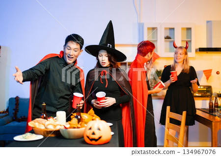 Young friends in Halloween costumes decorating pumpkin lanterns at a festive indoor party. Real people enjoying spooky vibes, red cups, snacks, and Halloween props. Young friends in Halloween costumes decorating pumpkin lanterns at a festive indoor party. Real people enjoying spooky vibes, red cups, snacks, and Halloween props. 134967076