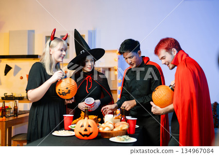 Young friends in Halloween costumes decorating pumpkin lanterns at a festive indoor party. Real people enjoying spooky vibes, red cups, snacks, and Halloween props. Young friends in Halloween costumes decorating pumpkin lanterns at a festive indoor party. Real people enjoying spooky vibes, red cups, snacks, and Halloween props. 134967084