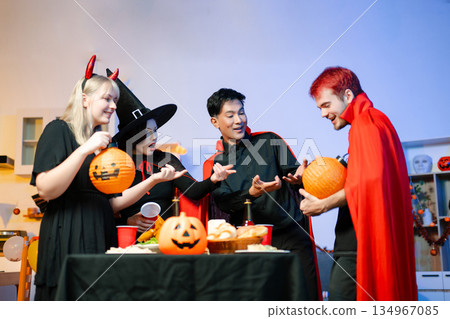 Young friends in Halloween costumes decorating pumpkin lanterns at a festive indoor party. Real people enjoying spooky vibes, red cups, snacks, and Halloween props. Young friends in Halloween costumes decorating pumpkin lanterns at a festive indoor party. Real people enjoying spooky vibes, red cups, snacks, and Halloween props. 134967085