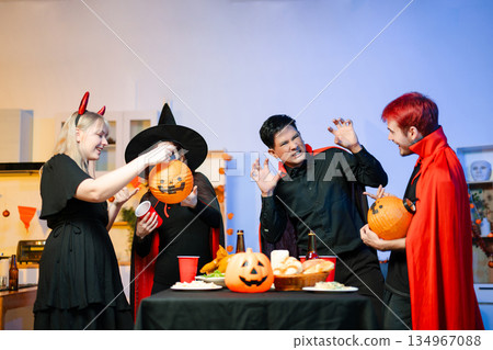 Group of friends in fun Halloween costumes laugh and play with spooky props, lanterns, and skull decor at a festive indoor party. Group of friends in fun Halloween costumes laugh and play with spooky props, lanterns, and skull decor at a festive indoor party. 134967088