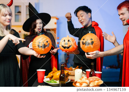 Group of friends in fun Halloween costumes laugh and play with spooky props, lanterns, and skull decor at a festive indoor party. Group of friends in fun Halloween costumes laugh and play with spooky props, lanterns, and skull decor at a festive indoor party. 134967139