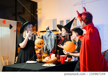 Group of friends in fun Halloween costumes laugh and play with spooky props, lanterns, and skull decor at a festive indoor party. Group of friends in fun Halloween costumes laugh and play with spooky props, lanterns, and skull decor at a festive indoor party. 134967144