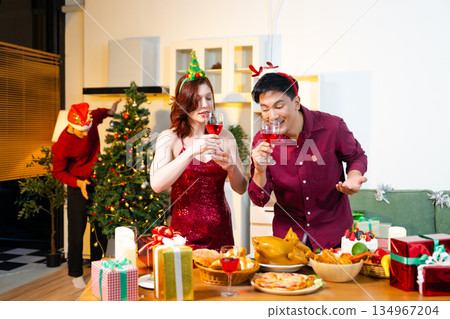 Group of cheerful friends celebrating Christmas with food, wine, and gifts around a festive decorated tree in warm joyful holiday atmosphere. 134967204