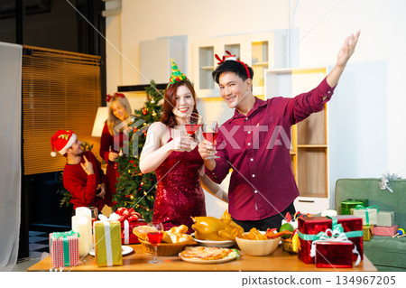 Group of cheerful friends celebrating Christmas with food, wine, and gifts around a festive decorated tree in warm joyful holiday atmosphere. 134967205