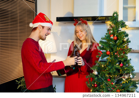 Smiling couple celebrating Christmas by exchanging gifts beside a decorated tree, sharing joy and festive holiday spirit in cozy indoor setting. 134967211