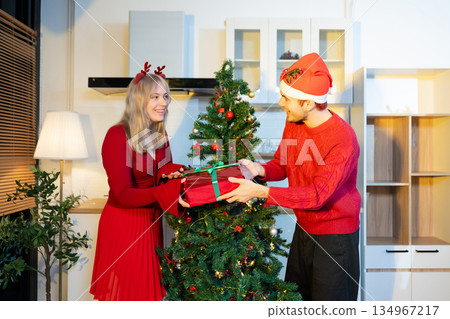 Happy couple exchanging gifts beside Christmas tree, sharing smiles, love, and festive holiday joy in a cozy indoor celebration atmosphere. 134967217