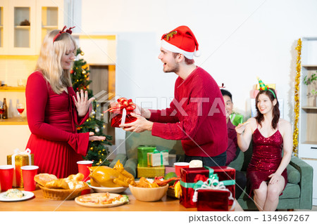 Friends celebrating Christmas together at a cozy home party with gift exchange, festive food and joyful holiday atmosphere. 134967267