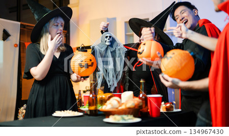 Group of friends in fun Halloween costumes laugh and play with spooky props, lanterns, and skull decor at a festive indoor party. 134967430