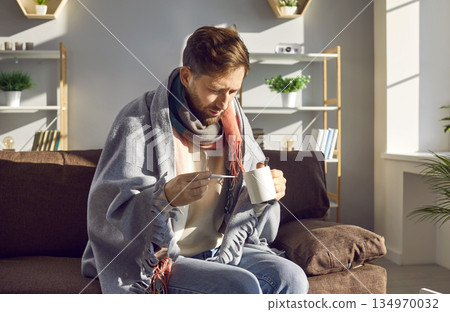 Sick young man sitting on sofa with thermometer wrapped in plaid 134970032