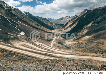 Winding dirt road through a mountain pass with rocks and snow-capped peaks Winding dirt road through a mountain pass with rocks and snow-capped peaks 134972381
