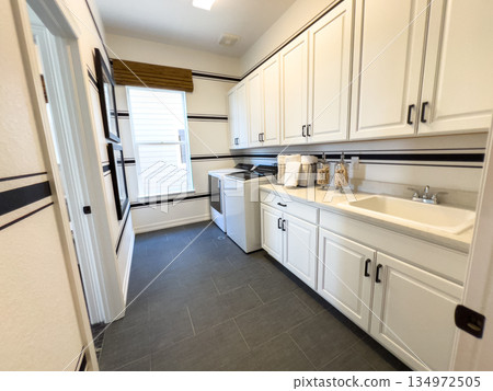 Well-lit laundry room with a large window, long countertop, sink, and white cabinetry. Decorative jars and black hardware add modern character to the space. 134972505