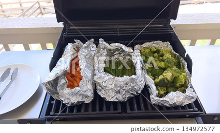 Fresh vegetables including carrots, broccoli, and asparagus are cooking in foil packets on an outdoor grill. The bright natural daylight enhances the texture and color of the vegetables in a casual 134972577