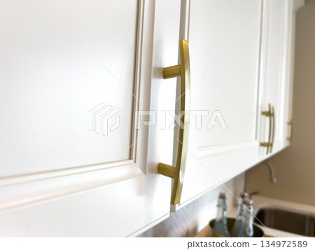 Detailed view of sleek gold handles on white kitchen cabinetry under bright light. The photo captures the elegant contrast and refined craftsmanship of the modern interior. 134972589