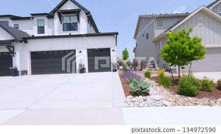 Two attached homes with dark garage doors are shown with a wide, clean concrete driveway. The front yards are landscaped with low shrubs and decorative rocks. 134972590